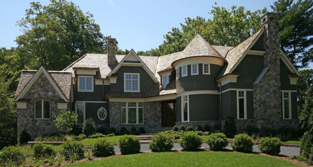 Elegant stone and shingle home with turret and lush landscaping in a sunny setting.