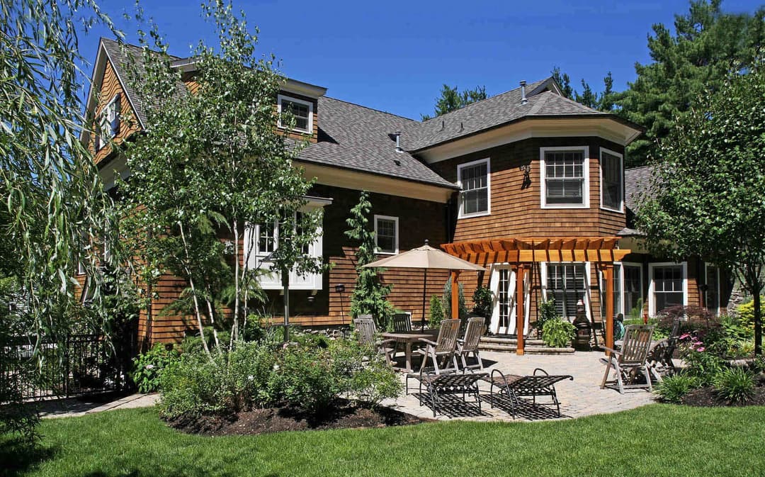 Charming wooden house with a patio, lush garden, and outdoor seating area in bright sunlight.