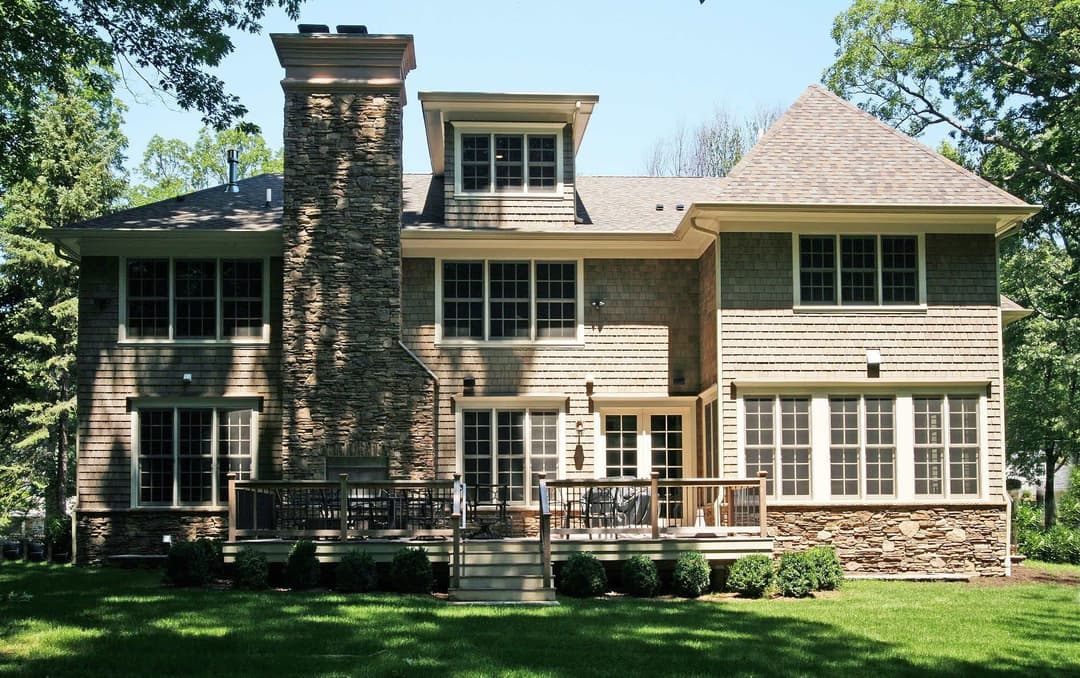 Modern two-story house with stone exterior, large windows, and a wooden deck in a landscaped yard.