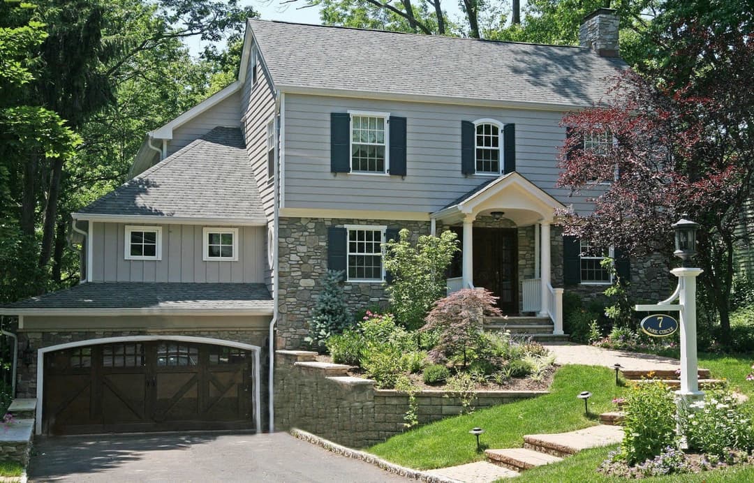 Charming gray stone house with landscaped garden and two-car garage, surrounded by greenery.